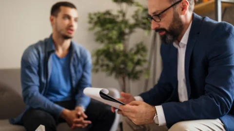 A middle-aged male clinician with glasses and a beard sits across from a younger male patient, reviewing a clipboard during an adult autism diagnosis assessment. REACH, Ohio