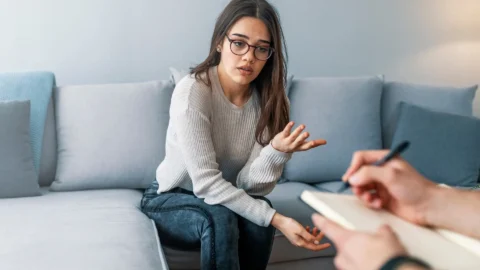 **Alt text:** `A young woman with glasses sits on a couch talking with a therapist taking notes, representing how REACH Behavioral Health uses an individual service plan to guide personalized therapy`