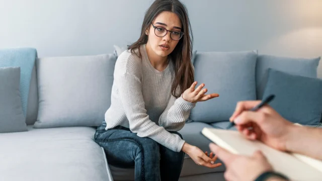 **Alt text:** `A young woman with glasses sits on a couch talking with a therapist taking notes, representing how REACH Behavioral Health uses an individual service plan to guide personalized therapy`