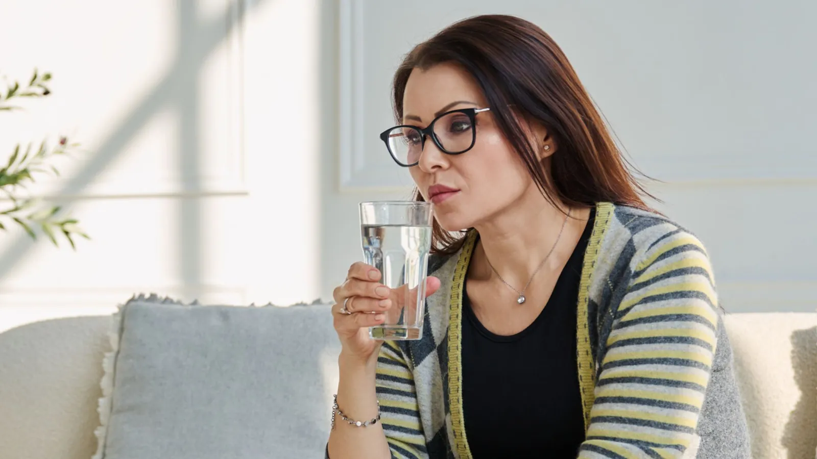 Woman with glasses sipping a glass of water while sitting on a sofa at home